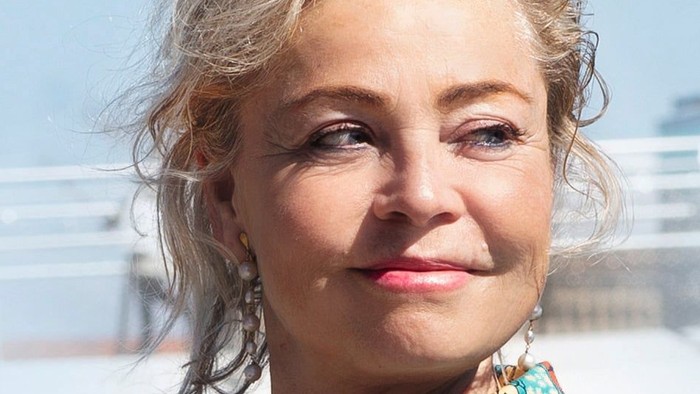 En kvinde med blondt hår i en blomstret bluse, der smiler udendørs ved en strandpromenade.