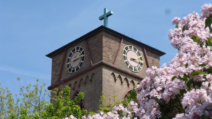 Ein Kirchturm mit Uhr und Kreuz, umgeben von blühenden Bäumen und einem klaren blauen Himmel