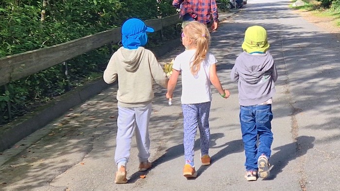 Drei Kinder gehen Hand in Hand auf einem gepflasterten Weg, umgeben von üppigem Grün und Bäumen.
