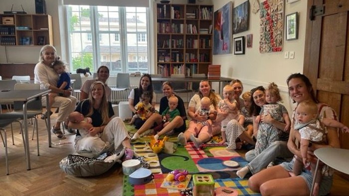 En gruppe af voksne og børn sidder på gulvmatte i et rum med bogshelve og billeder på væggen.