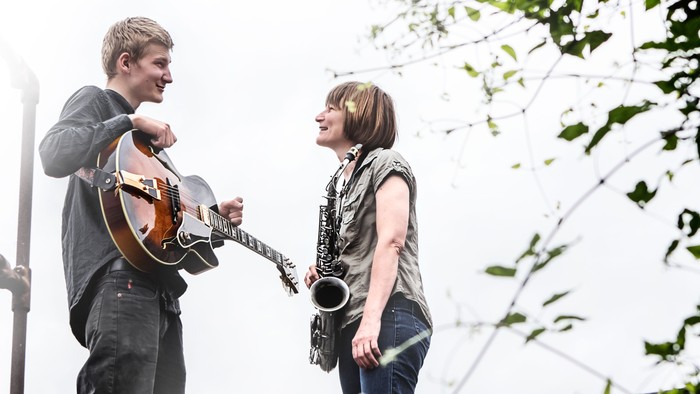 To musikere, en guitarist og en saxofonist, smiler mens de optræder udendørs.
