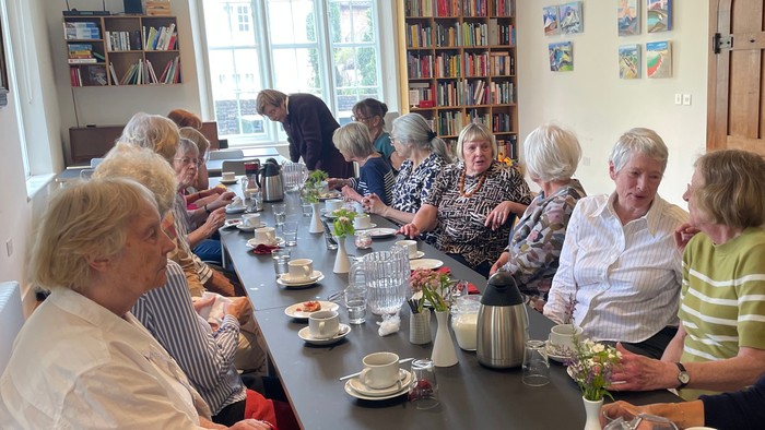 En gruppe nyder te og samtale omkring et langt spisebord i et hyggeligt, bogfyldt rum.