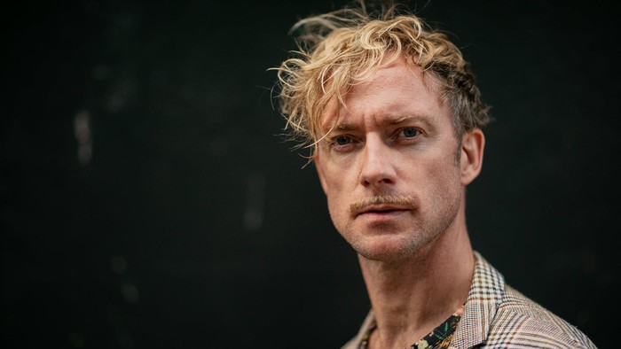 En mand med krøllet hår og overskæg bærer en ternet blazer over en farverig skjorte på en mørk baggrund.