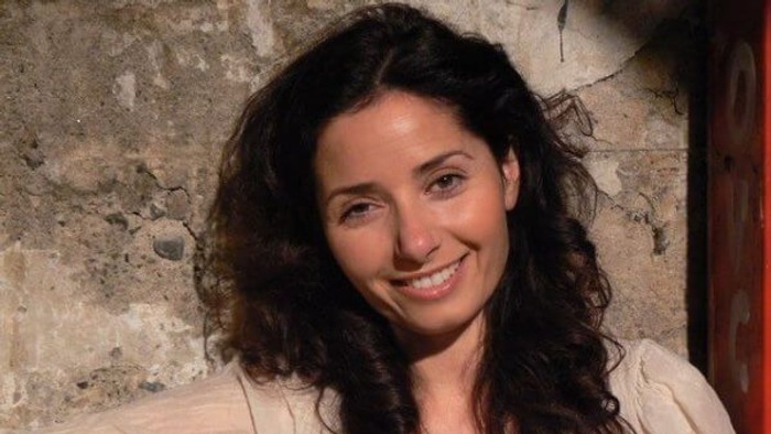 En smilende kvinde med langt mørkt hår iført en lys bluse foran en rustik stenmur.