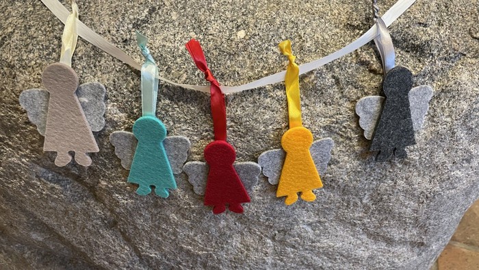 Farverige filt-engeldekorationer med vinger, der hænger på en snor mod en tekstureret baggrund.