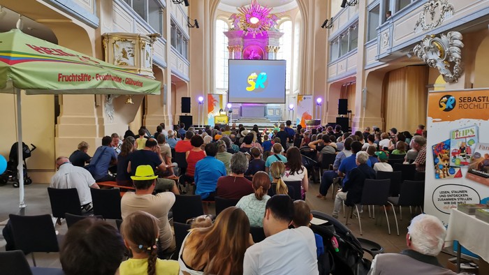 Große Menschenmenge in einem prächtigen Saal, viele sitzen, einige stehen, große Bildschirme und bunte Dekorationen