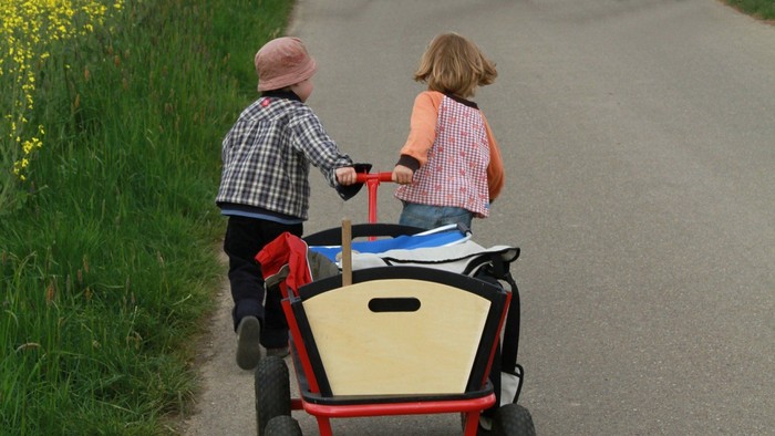 Zwei Kinder fahren mit einem roten Dreirad auf einer Straße.