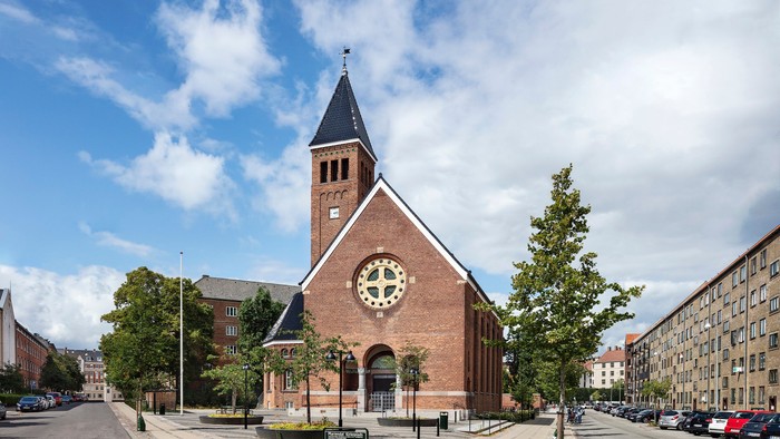 Murstenskirke med højt spir og ur, omgivet af træer og bygninger.