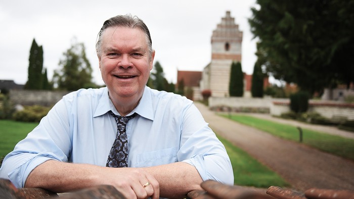 En smilende mand i en lyseblå skjorte og slips poser udendørs nær en historisk stenbygning og kirkegård.
