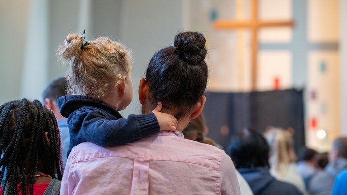 Eine Frau mit einem Kind im Arm steht in einer Kirche und blickt mit anderen zum Altar.