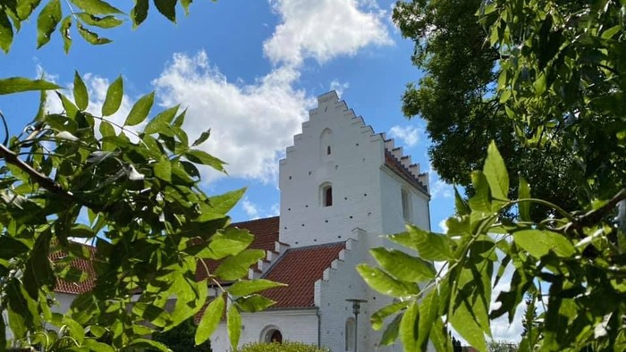 Et hvidt kirkebygning med røde tag og en tårn, omgivet af grønne træer og buske