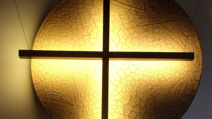 Eine leuchtende, kreisförmige Lichtinstallation in Kreuzform, die an einer strukturierten Wand montiert ist.