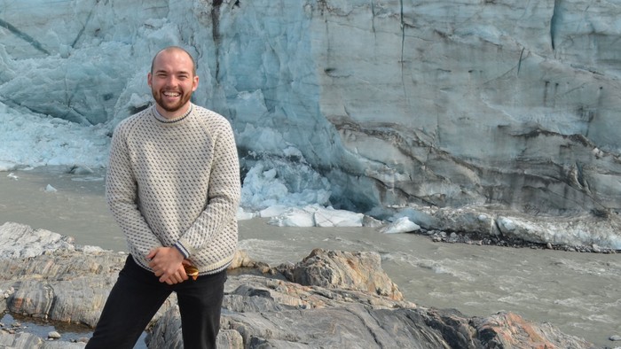 En person står nær en enorm gletsjer med ujævne isformationer og en stenet forgrund.