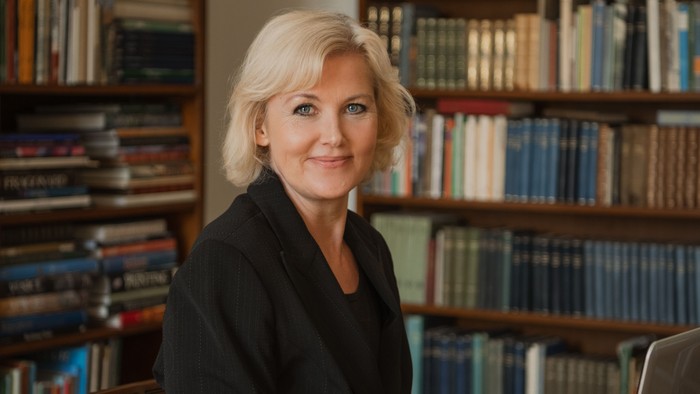 Professionel kvinde, der arbejder ved et skrivebord omgivet af bøger, og bruger en bærbar computer og en åben bog.