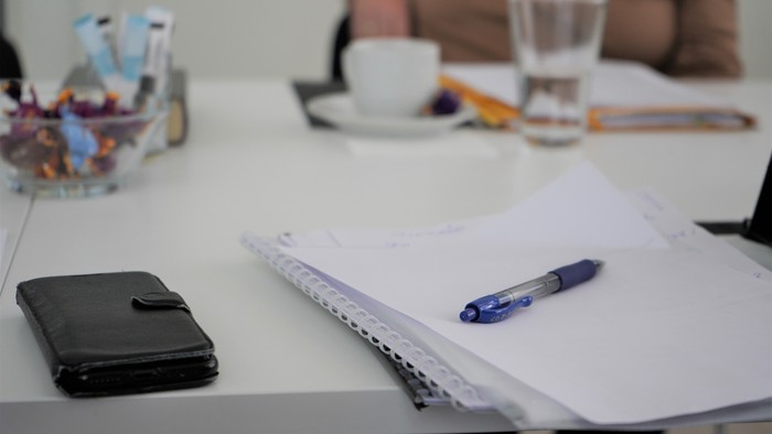 Et ryddeligt arbejdsområde med notesbog, kuglepen, pung, kaffe og et glas vand på et bord.
