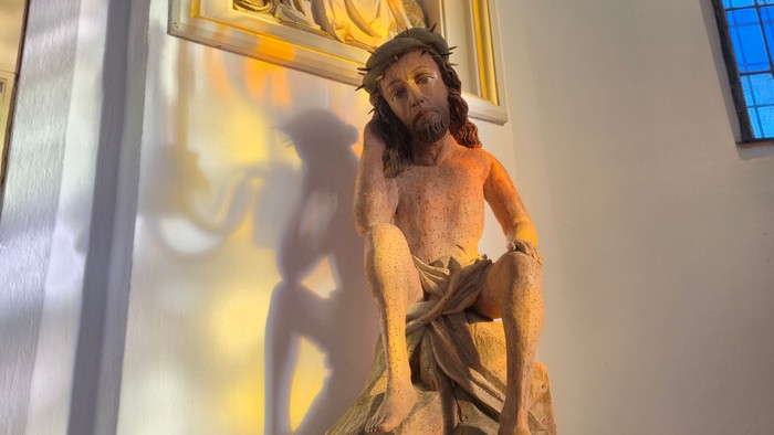 Eine Statue von Christus, der das Kreuz trägt, auf einem Sockel mit religiösen Kunstwerken darüber.