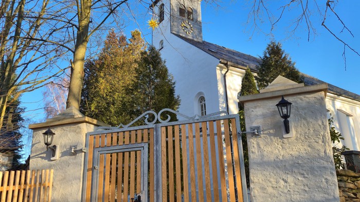 Eine friedvolle Kirche mit hohem Kirchturm, gesehen durch ein kunstvolles, holzgerahmtes Tor und eine Steinmauer.