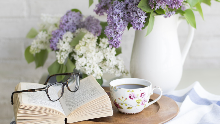 Eine gemütliche Szene mit einem offenen Buch, einer Brille, einer blumigen Teetasse und blühenden Fliederzweigen in einer Vase.