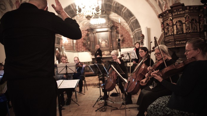 En dirigent leder et strygeorkester, der optræder i en historisk, svagt oplyst kirke.