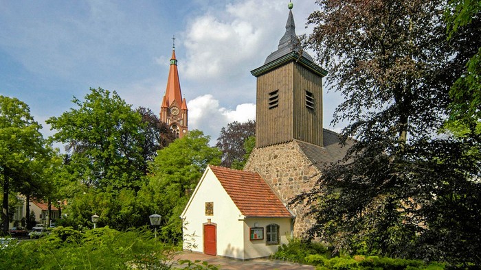 Kirche mit Uhrturm und kleinerem Gebäude davor, umgeben von Bäumen und Grünflächen.