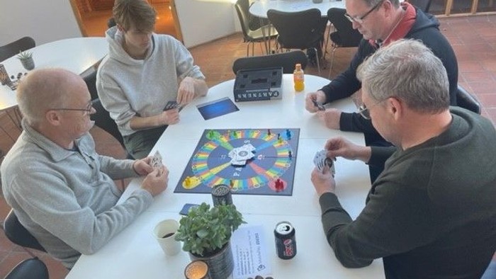 En gruppe mennesker spiller brætspil på et rundt bord i et rum med trægolv og store vinduer.