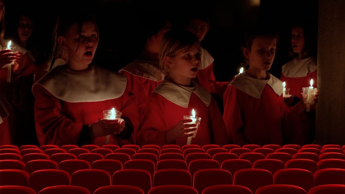 Kinder in roten Roben halten Kerzen, während sie in einem leeren Theater singen.