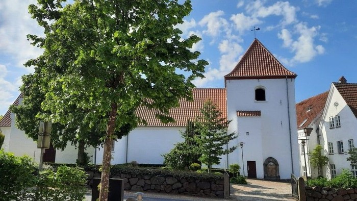 Et hvidt kirkebygningskompleks med røde tag og træer foran