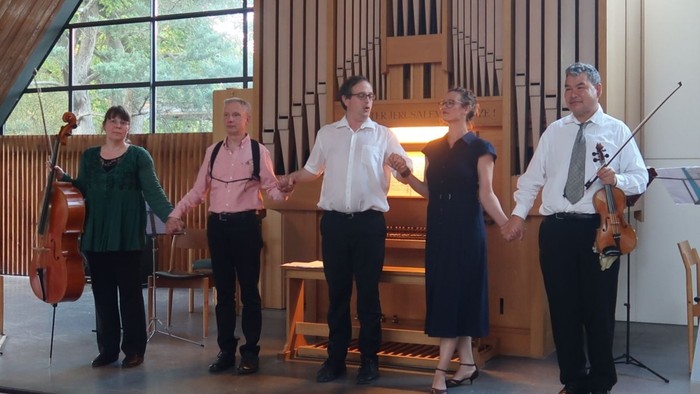 Ein kleines Ensemble spielt in einem geräumigen Saal mit einer großen Orgel, und die Musiker halten sich an den Händen.