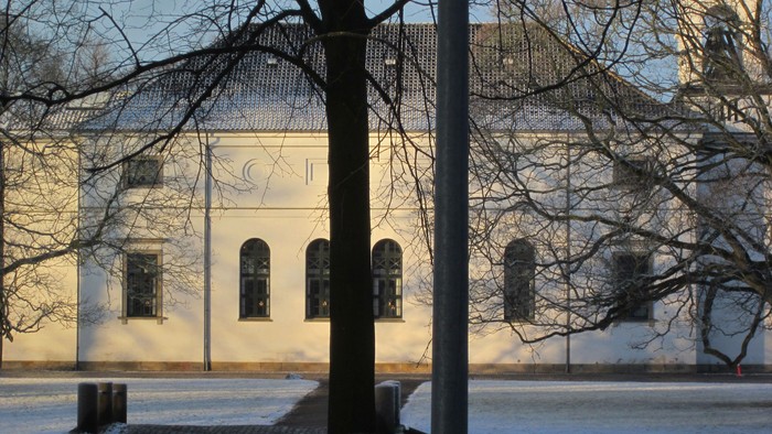 En snedækket kirke med to vinduer og et tårn bagved en træ.