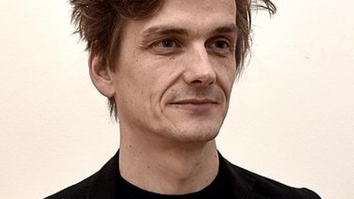 En person med kort brunt hår iført en sort jakke på en ensfarvet baggrund.