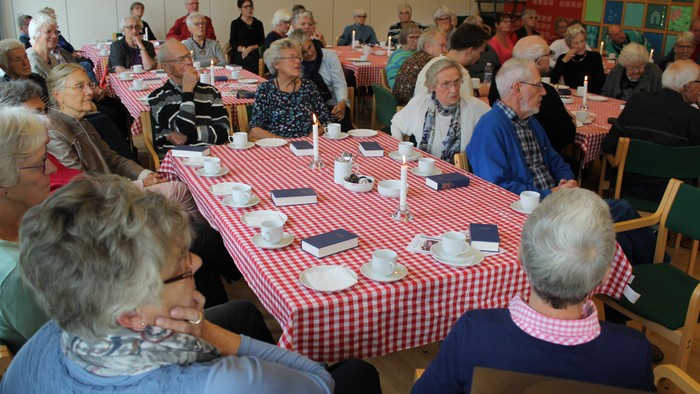 En gruppe ældre samles ved lange bord med røde og hvide tæpper, der er dækket med tebeklædning og servies. De sidder og taler med hinanden i et rum med grønne stoler.