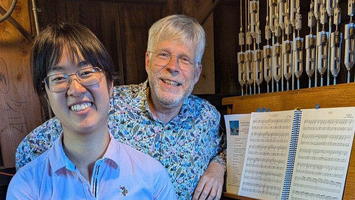 To personer smiler ved siden af et stort træmusikinstrument med synlige tangenter og noder.