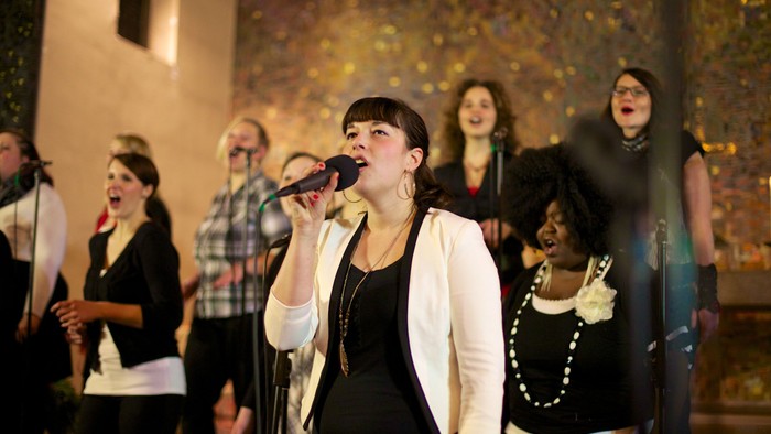 Eine Frau singt in ein Mikrofon, umgeben von anderen Sängerinnen.