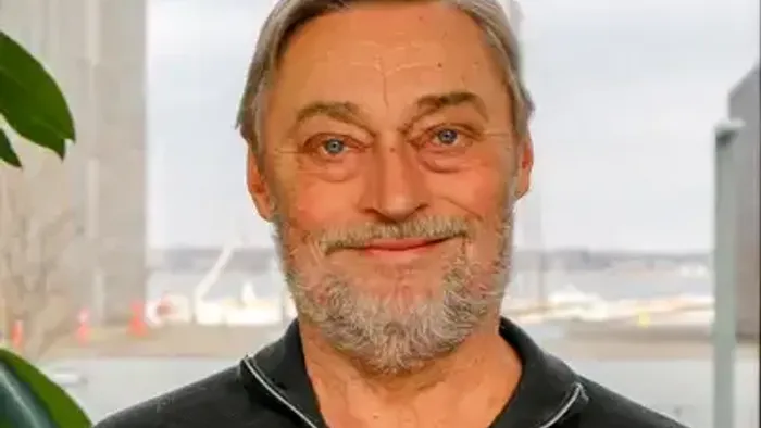 En smilende mand med skæg, der står udendørs nær en havnefront med industrielle bygninger i baggrunden.