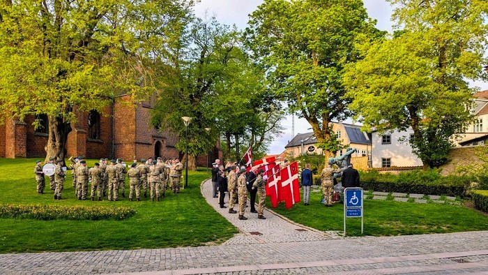 En gruppe uniformerede personer står i linje med flag på en græsplads ved et historisk bygning.