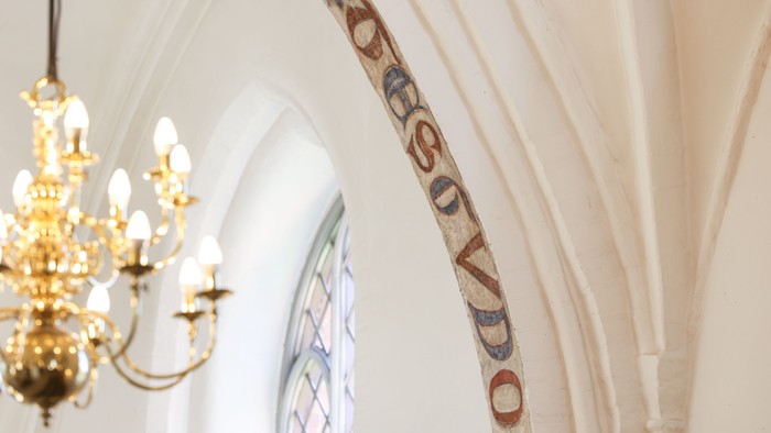 En belysning hænger fra loftet i en kirke med et bogstaveligt maleri på væggen.