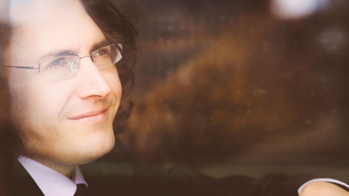 Ein Mann im Anzug mit Krawatte lehnt lässig an einem Geländer und lächelt leicht im Freien.