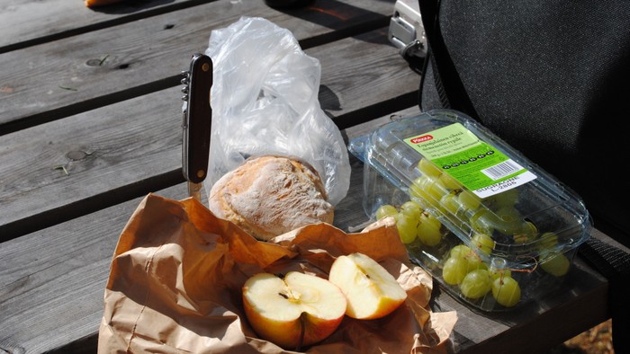 Auf einem Holztisch liegen ein belegtes Brötchen, ein aufgeschnittener Apfel und eine Packung Weintrauben. Daneben liegen eine Brottüte, eine durchsichtige Verpackung und ein Taschenmesser.