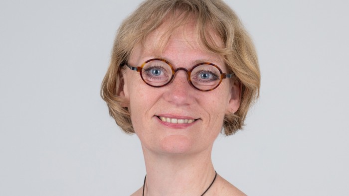 En smilende kvinde med blondt hår, iført runde briller og en mørk off-shoulder top.