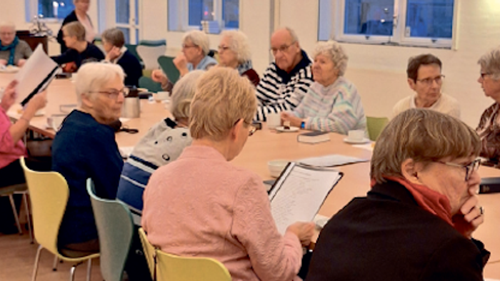 En gruppe ældre personer sidder i en kreds og deltager i en aktivitet i et rum med store vinduer.