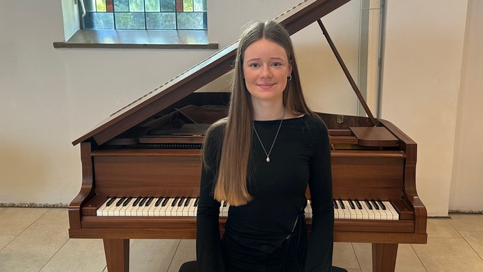 Eine junge Frau sitzt an einem aufrechten Klavier mit einem fröhlichen Gesichtsausdruck und trägt ein schwarzes Oberteil.