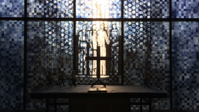 Ein Raum mit einem Tisch und einem Kreuz davor, vor einem großen Fenster mit buntem Glas.