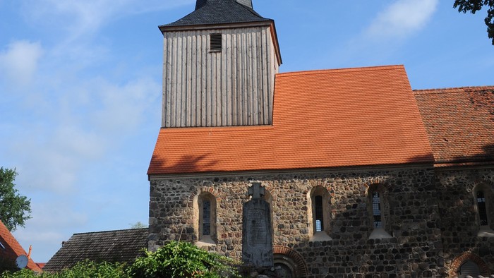 Kirchturm mit spitzem Dach und Uhr, umgeben von Steinmauer und Büschen