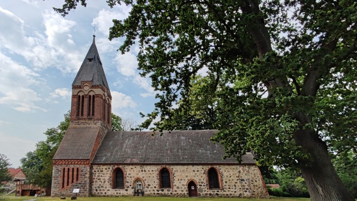 Das Bild zeigt eine alte Kirche mit hohem Turm und Steinmauern, umgeben von Bäumen und grünem Gras.
