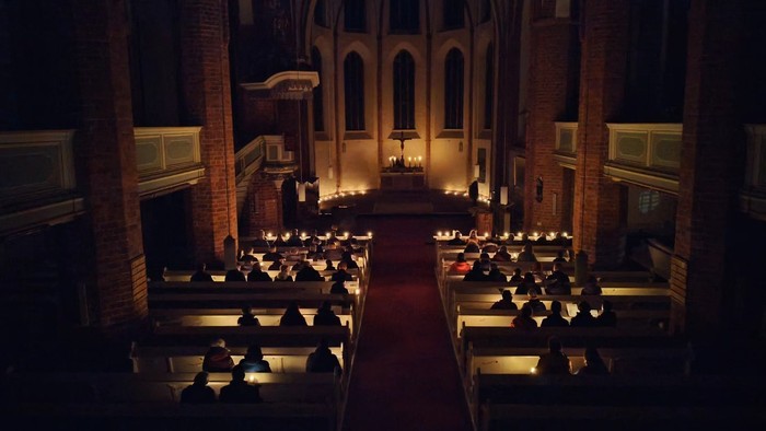 Ein Kircheninneres bei einer Kerzenzeremonie, mit Menschen in Reihen sitzend und einem Mittelgang mit brennenden Kerzen.