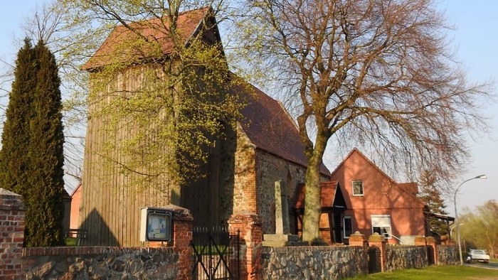 Ein rotes Backsteingebäude mit spitzem Dach und hohem Tor, umgeben von Bäumen und einem Laternenpfahl.