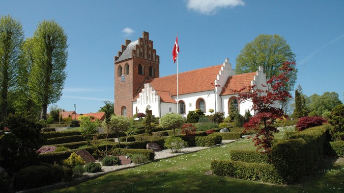 En smuk kirke med røde tag og en flagstang foran, omgivet af en grøn og blomstrende have