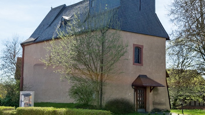 Kirche mit hohem Turm und Kreuz, umgeben von Bäumen und Gras.