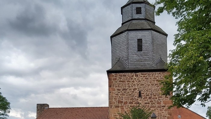 Historische Steinkirche mit modernem Glasanbau, umgeben von Grünflächen und vor bewölktem Himmel gelegen.