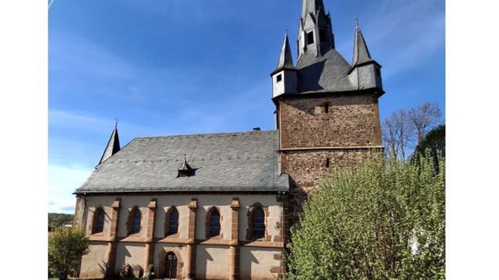 Kirche mit hohem Turmspitz und Rundbogenfenstern, umgeben von grünem Gras und Sträuchern.